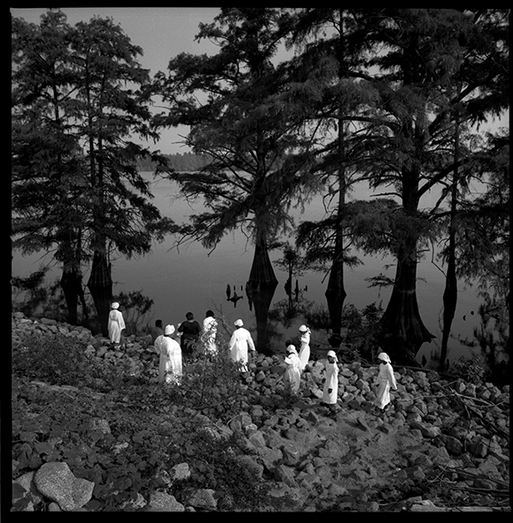 Baptism II Moon Lake, MS 1997. A group of baptism candidates make their way carefully over loose stones to the edge of Moon Lake, an oxbow of the Mississippi near Lula, MS. Small churches in the Lula/Friars Point area gather in the fall to baptize candidates in the lake just as they have for over one hundred years. The great migration of blacks from the Delta in the 1940’s and 50’s resulted in a dramatic decline in the Delta population, impacting church congregations to the point that many churches were abandoned. Today’s Delta churches now pool their resources to help keep tradition alive. Baptisms are usually attended by members of 3 to 6 different congregations.